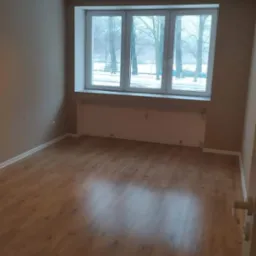 Pusty pokój po remoncie z nową podłogą z paneli laminowanych o ciepłym odcieniu drewna, świeżo pomalowanymi ścianami w kolorze beżowym i widokiem na park przez okno.