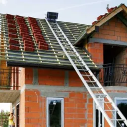 Budowa dachu z czerwonej dachówki na dwupiętrowym domu z cegły w Oświęcimiu, widoczna drabina transportująca materiały na górę.