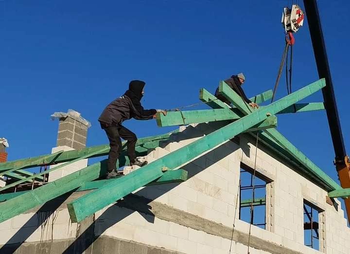 Dwóch dekarzy montuje zielone drewniane krokwie na dachu domu z białego pustaka, z widocznym kominem i dźwigiem w tle, przy słonecznej pogodzie.