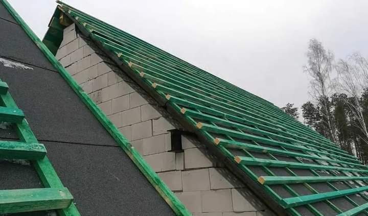 Konstrukcja dachu z zielonymi łatami na murze z białych bloczków, częściowo pokryta czarną membraną dachową, widok na las w tle.