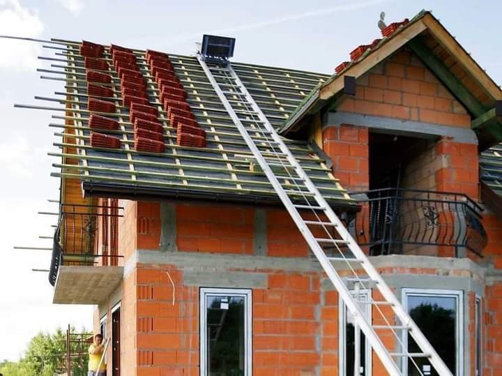 Budowa dachu z czerwonej dachówki na dwupiętrowym domu z cegły w Oświęcimiu, widoczna drabina transportująca materiały na górę.