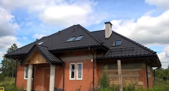 Dom w trakcie budowy z nowym, czarnym dachem z dachówki z oknami połaciowymi i białym kominem. Część elewacji z cegły, część zabezpieczona deskami. Widoczne rynny.