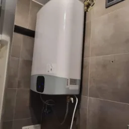 Bojler elektryczny Ariston zamontowany na szarej, kafelkowanej ścianie w łazience, widoczne podłączenia hydrauliczne i przewód zasilający.