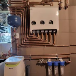 Kompleksowa instalacja kotła gazowego z miedzianymi rurami i filtrami wody na ścianie z szarych płytek, widoczny zbiornik wyrównawczy i zmiękczacz wody USTM.