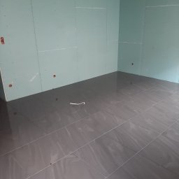Usługi ogólnobudowlane Bartłomiej Jakubiak - Świeżo położone, błyszczące, szare płytki podłogowe w pustym pomieszczeniu z zamontowanymi płytami gipsowo-kartonowymi na ścianach, widoczne otwory na gniazdka elektryczne.
