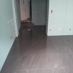 Usługi ogólnobudowlane Bartłomiej Jakubiak - Wnętrze w trakcie remontu: szare płytki podłogowe z delikatnym wzorem ułożone w nowym pomieszczeniu, ściany wykończone płytami gipsowo-kartonowymi, widoczne przygotowane otwory na gniazdka...