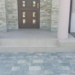 Usługi ogólnobudowlane Bartłomiej Jakubiak - Wykończone płytkami schody wejściowe z dwoma kolumnami, prowadzące do brązowych drzwi z dekoracyjnymi szybkami, z widoczną elewacją z kamienia dekoracyjnego i częściowo ułożoną kostką brukową...