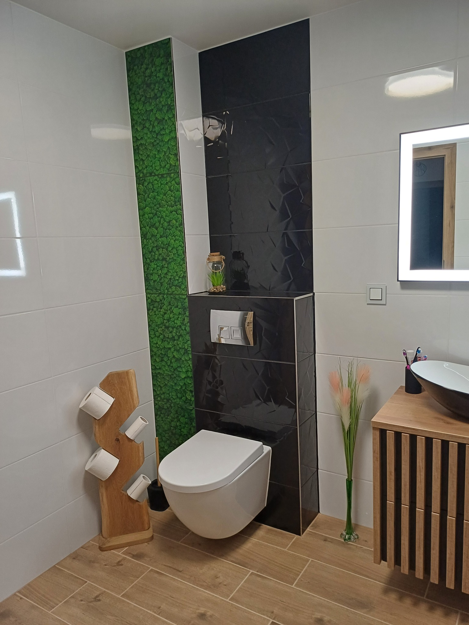 Nowoczesna łazienka z biało-czarnymi płytkami, panelem z mchu, podwieszaną toaletą i drewnianymi akcentami, takimi jak szafka pod umywalkę i stojak na papier toaletowy z surowego drewna.