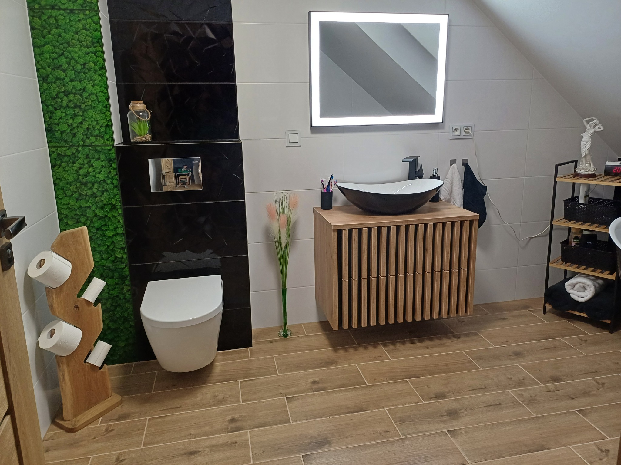 Nowoczesna łazienka na poddaszu z drewnopodobną podłogą, wiszącą toaletą, umywalką na szafce z lamelami, podświetlanym lustrem, czarnymi płytkami z fakturą i panelem z mchu.