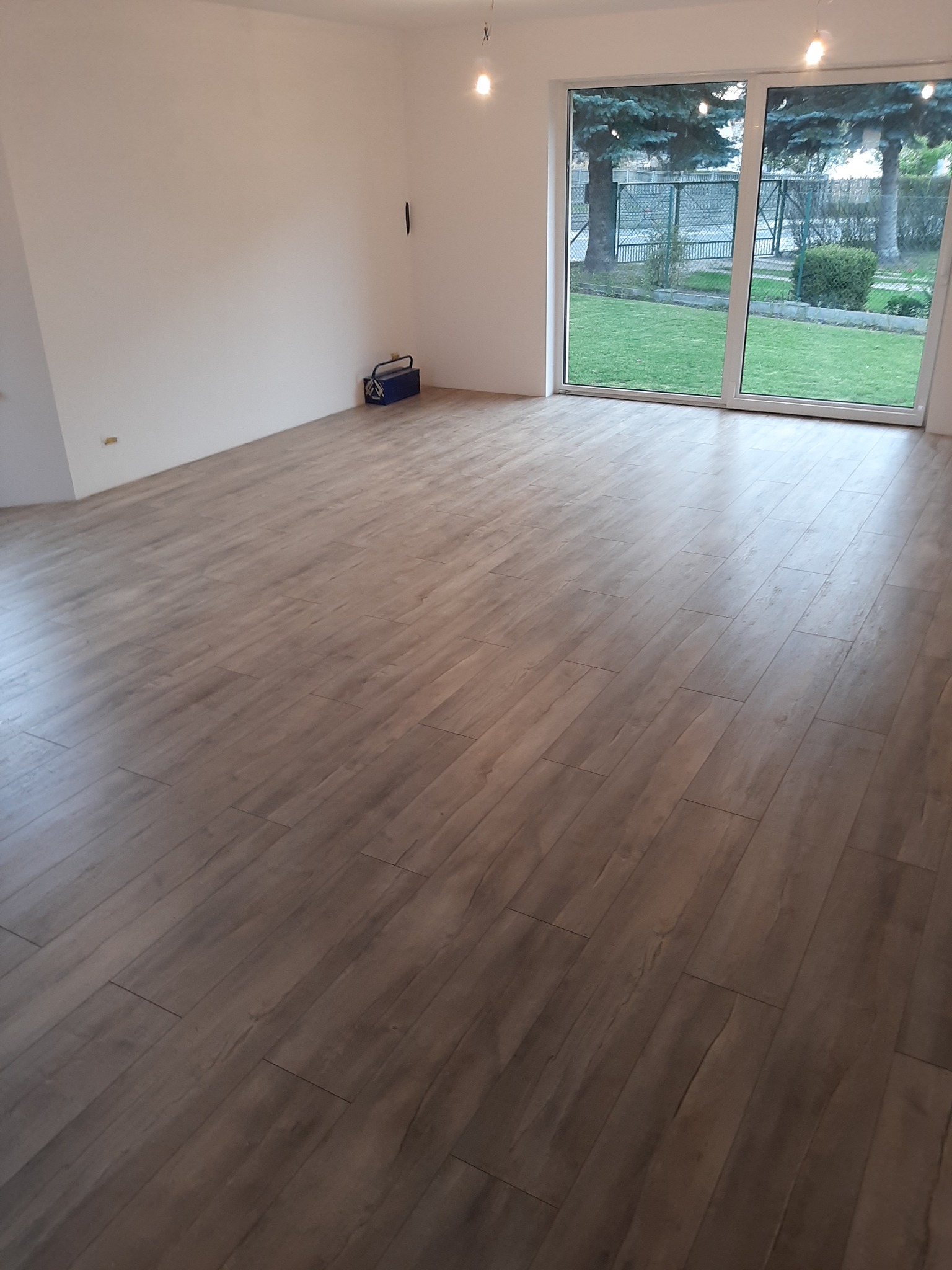 Jasny pokój z nowo ułożonymi panelami podłogowymi imitującymi drewno, widok na ogród przez duże okno przesuwne, na ścianie widoczne gniazdka elektryczne i skrzynka narzędziowa.