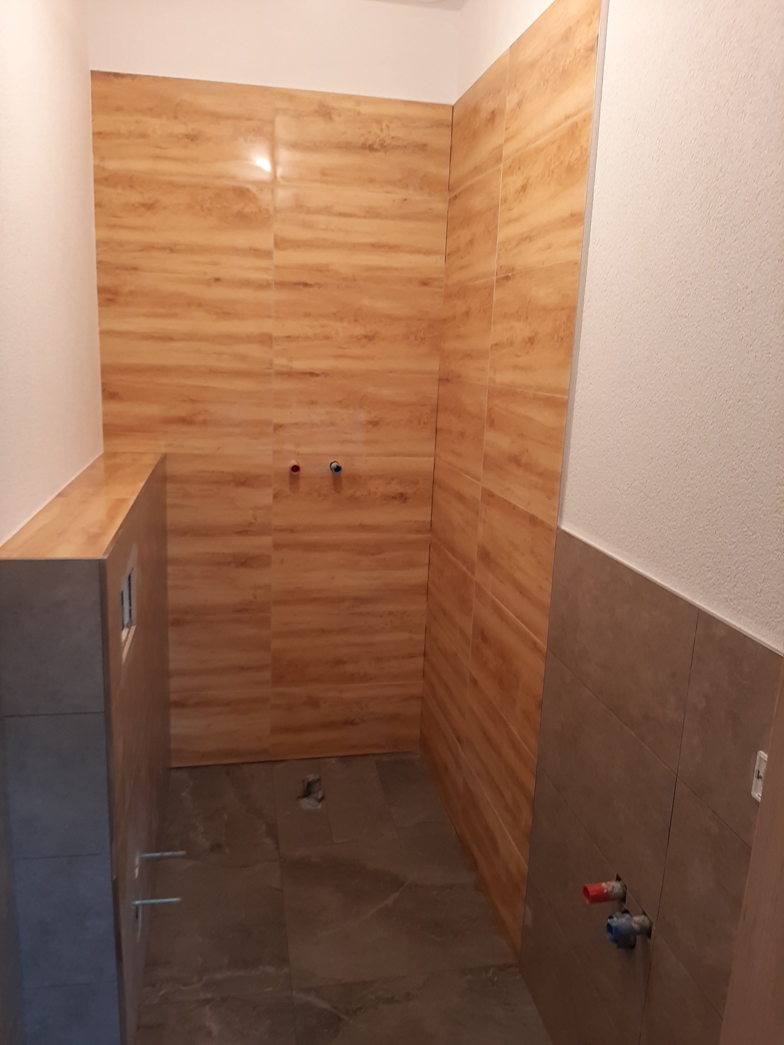Fragment łazienki w trakcie wykańczania: brązowe płytki imitujące drewno na ścianach i podłodze, widoczne wystające rury instalacji wodnej i kanalizacyjnej, częściowo położona glazura.
