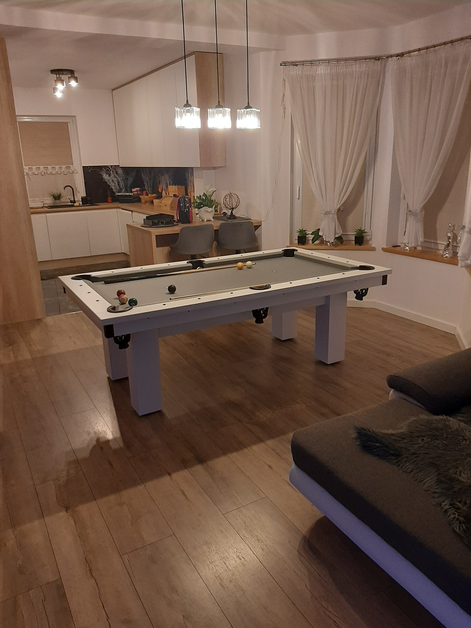 Stół bilardowy z szarym suknem w jasnym, nowoczesnym wnętrzu z drewnianą podłogą i kuchnią w tle, oświetlony wiszącymi lampami.