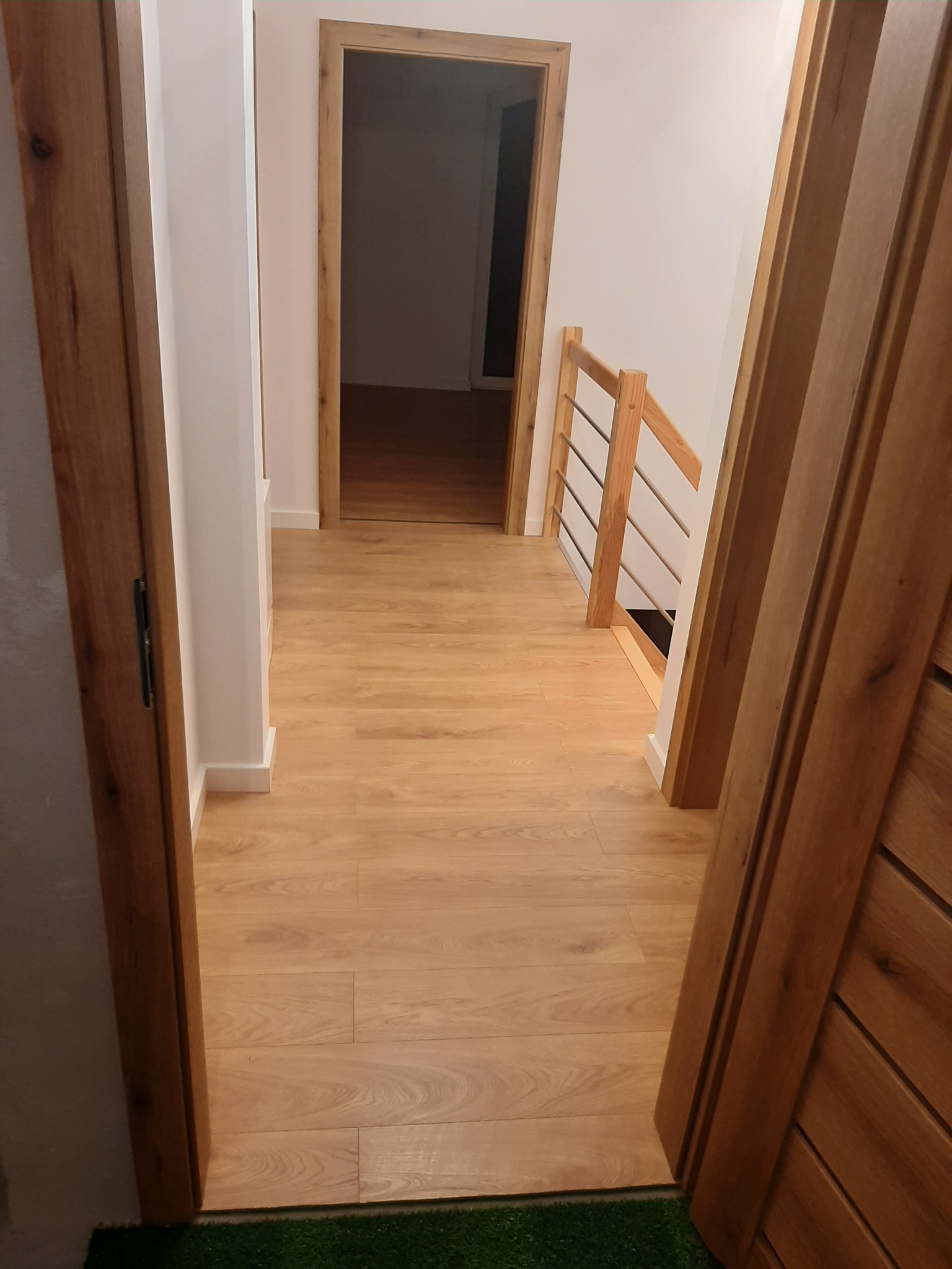 Jasne panele podłogowe w korytarzu z drewnianą balustradą i ościeżnicami, widok zza otwartych drzwi z zieloną wycieraczką.