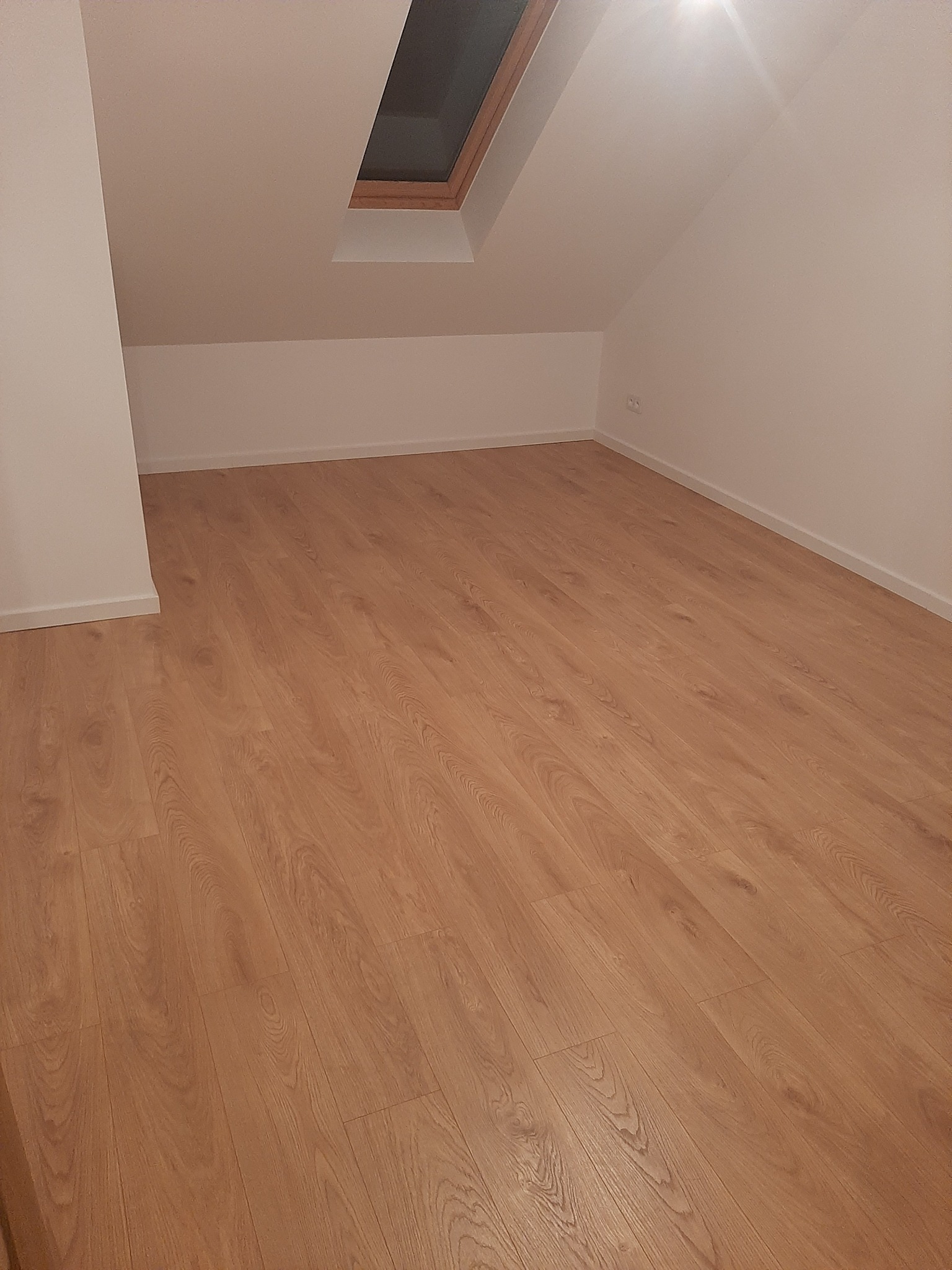 Jasne panele podłogowe w pokoju na poddaszu z oknem dachowym, świeżo po ułożeniu, widoczna listwa przypodłogowa.