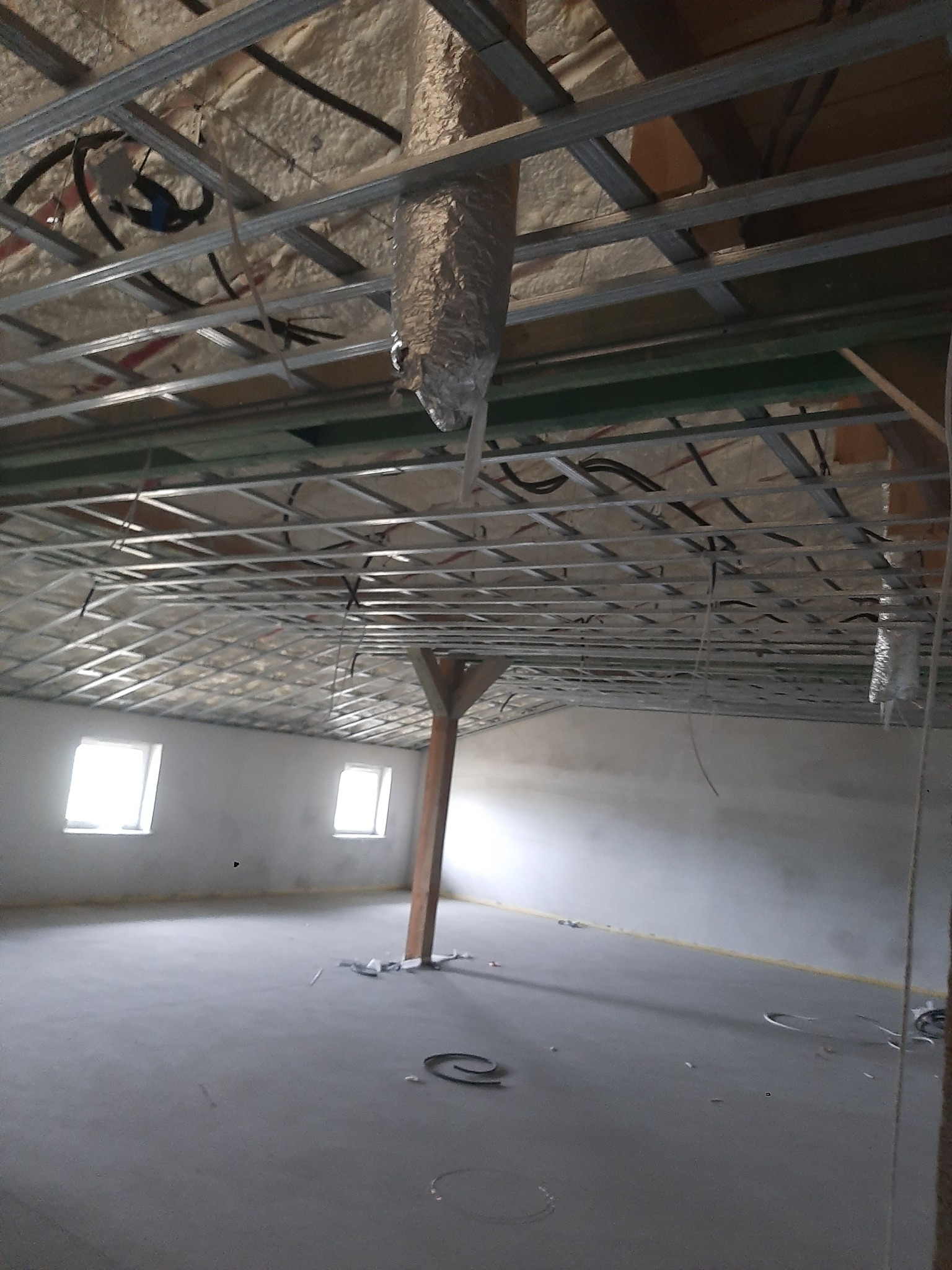 Surowy stan poddasza z widoczną izolacją piankową pod dachem, metalową konstrukcją sufitu podwieszanego i betonową wylewką podłogi, oświetlonego przez dwa okna.