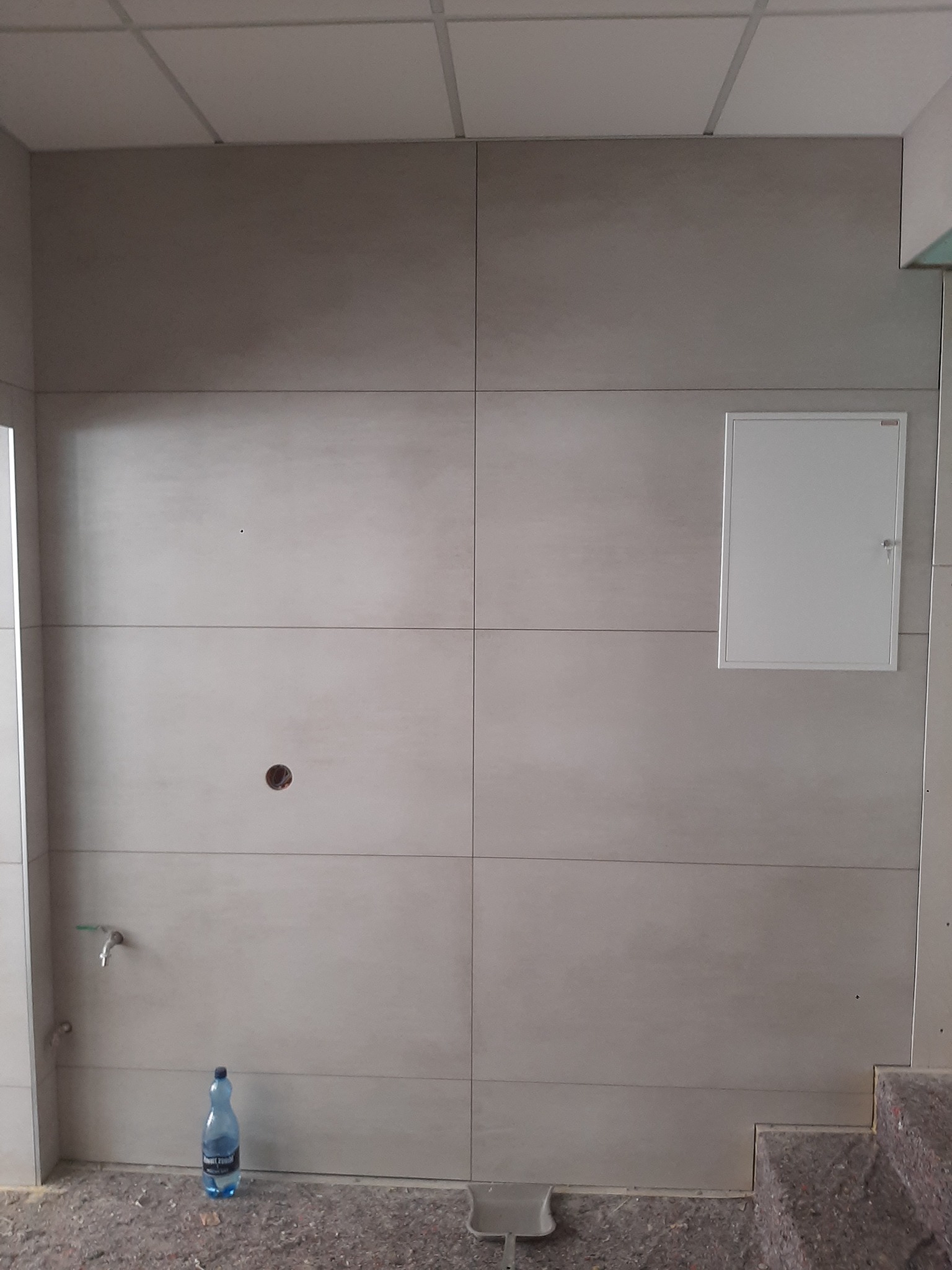 Ściana łazienki wyłożona jasnymi, wielkoformatowymi płytkami. Widoczny otwór na instalację, kran, butelka wody na podłodze i klapa rewizyjna. Podłoga zabezpieczona folią malarską.