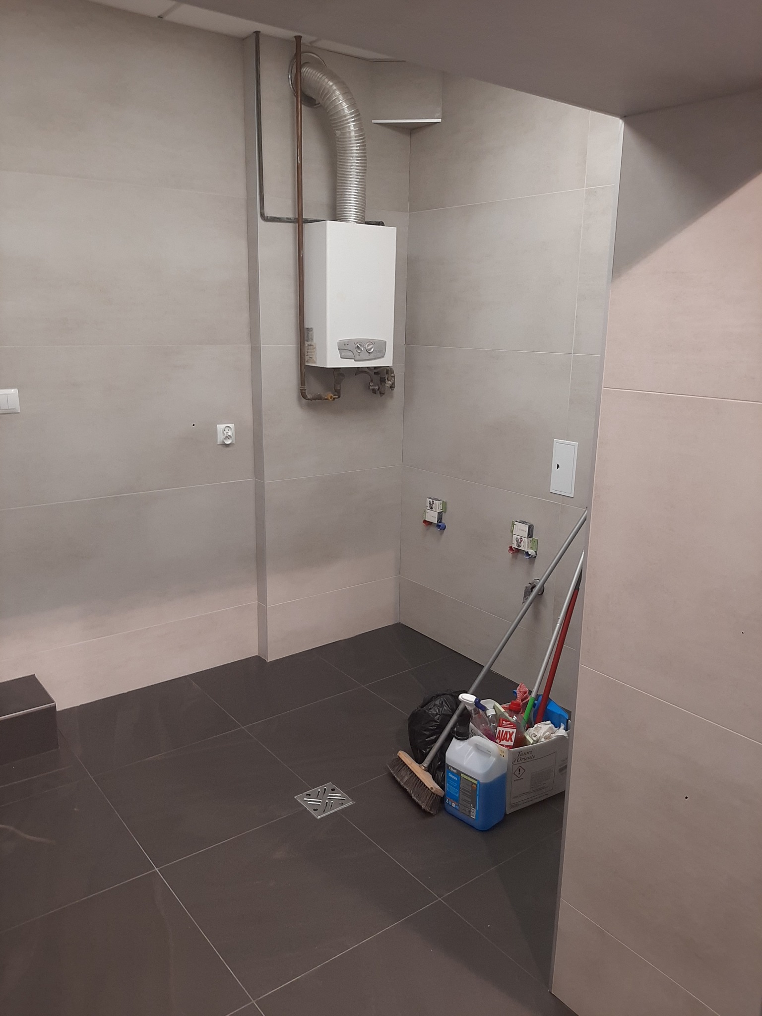 Wnętrze łazienki w trakcie prac wykończeniowych, widoczny piec gazowy z podłączeniem, ciemna podłoga z odpływem liniowym oraz wiadro ze środkami czystości i mopami.
