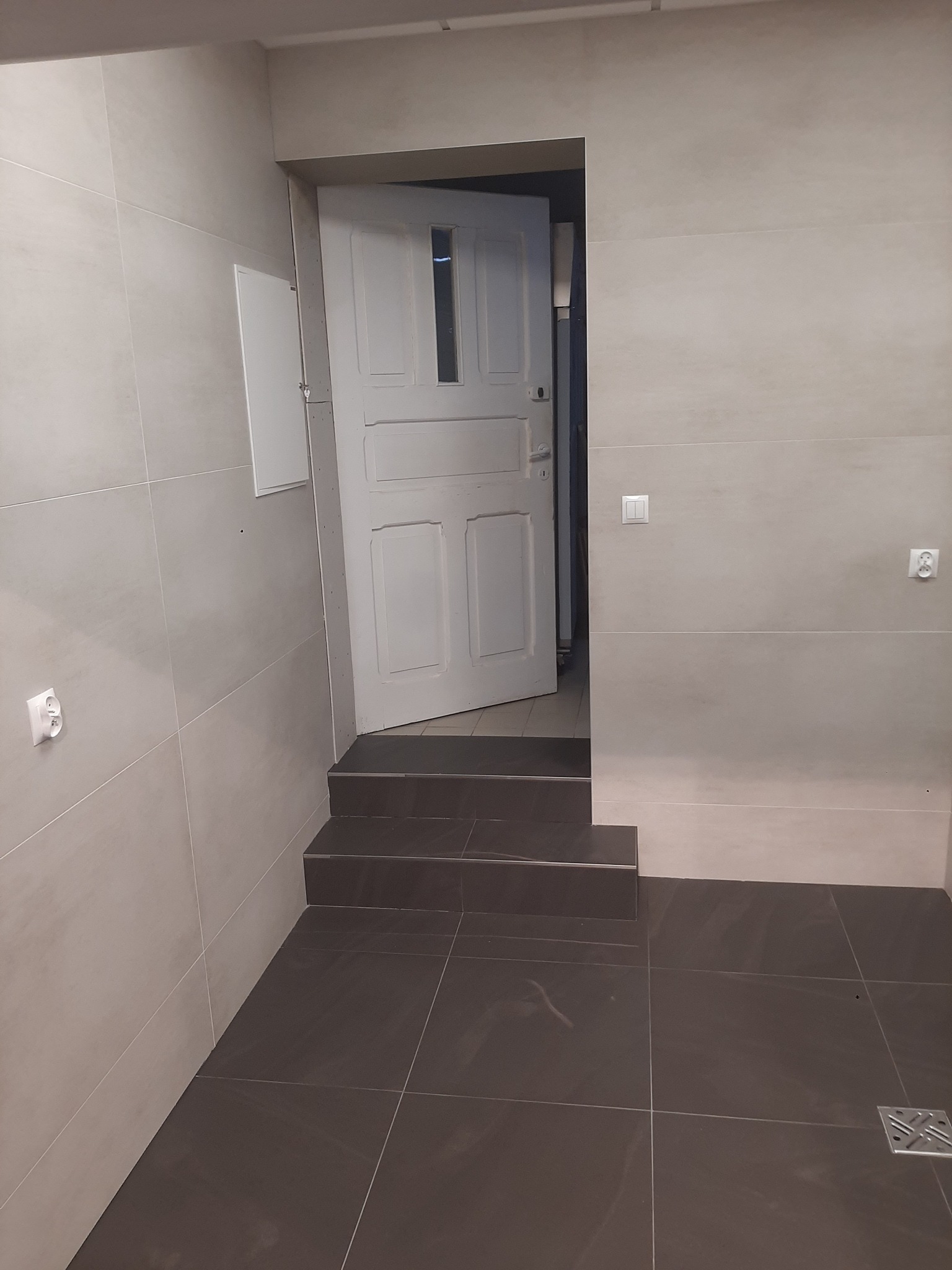 Wykończona łazienka z szarymi płytkami na ścianach i podłodze, widoczne trzy stopnie przed otwartymi białymi drzwiami oraz kratka odpływowa w rogu podłogi.