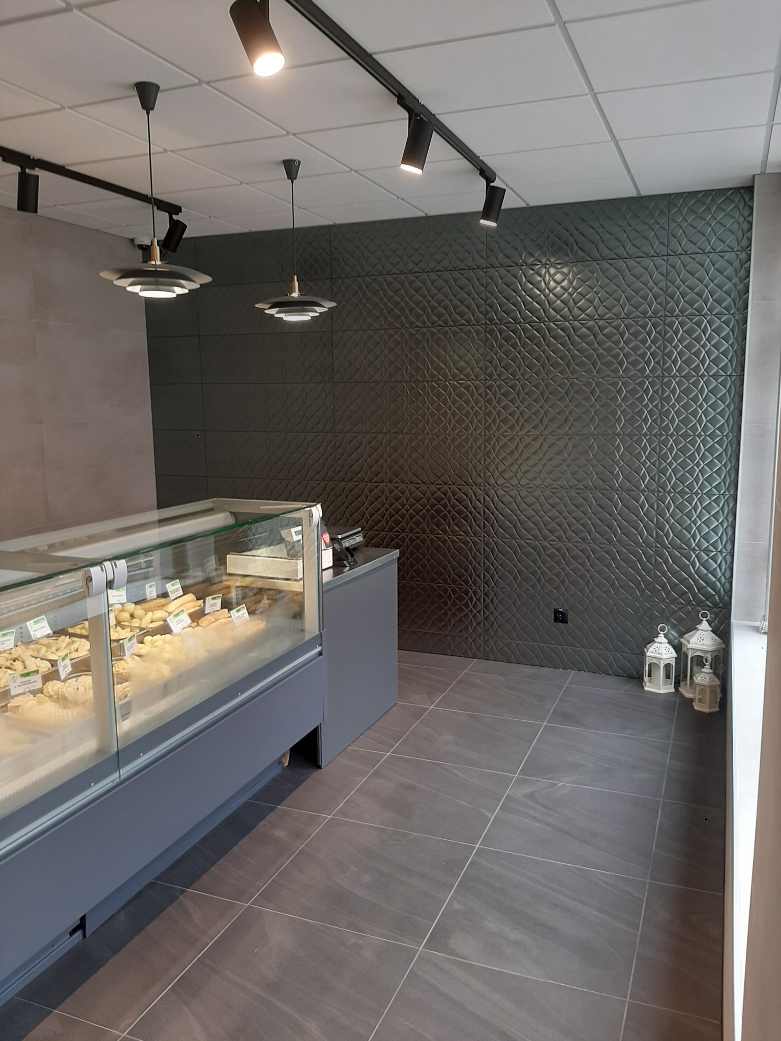 Wnętrze lokalu usługowego z szarymi płytkami podłogowymi imitującymi kamień, dekoracyjną ścianą z ciemnoszarych płytek 3D, oświetleniem punktowym i wiszącymi lampami oraz witryną chłodniczą...