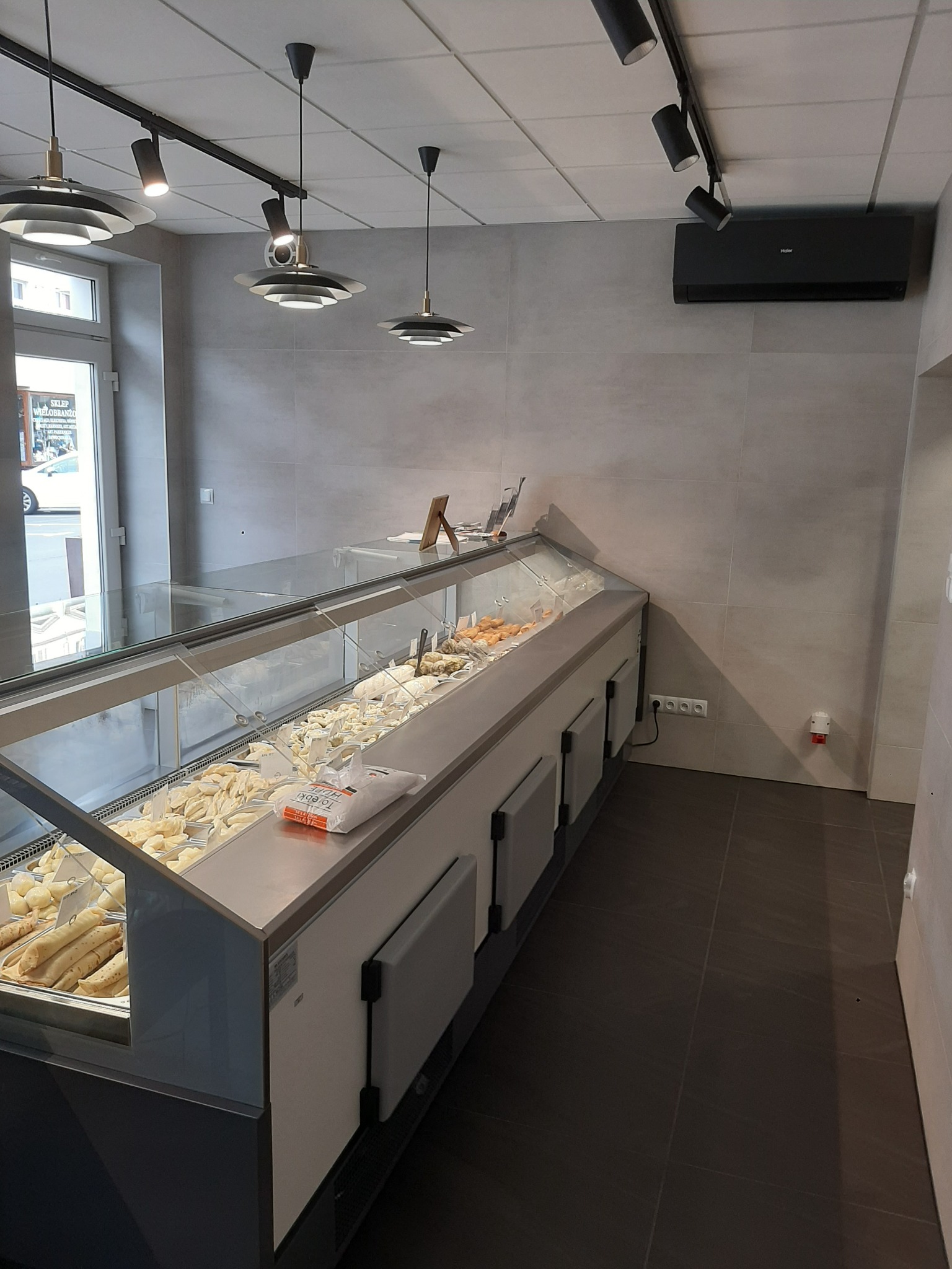 Wnętrze sklepu spożywczego po remoncie, z przeszkloną ladą chłodniczą wypełnioną pierogami i naleśnikami, nowoczesne oświetlenie sufitowe, szare płytki ścienne i ciemna podłoga.