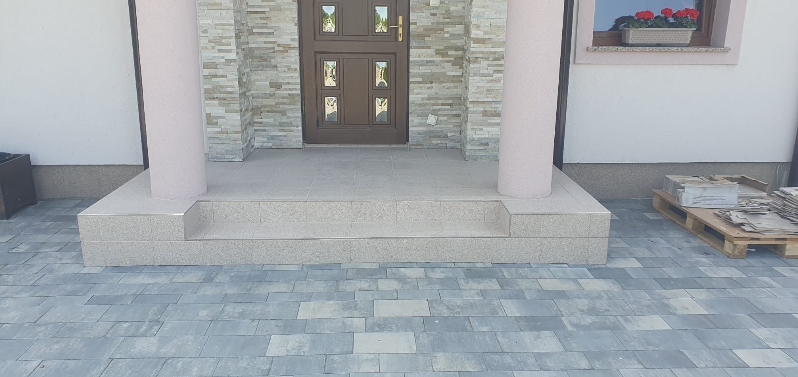 Wykończone płytkami schody wejściowe z dwoma kolumnami, prowadzące do brązowych drzwi z dekoracyjnymi szybkami, z widoczną elewacją z kamienia dekoracyjnego i częściowo ułożoną kostką brukową...