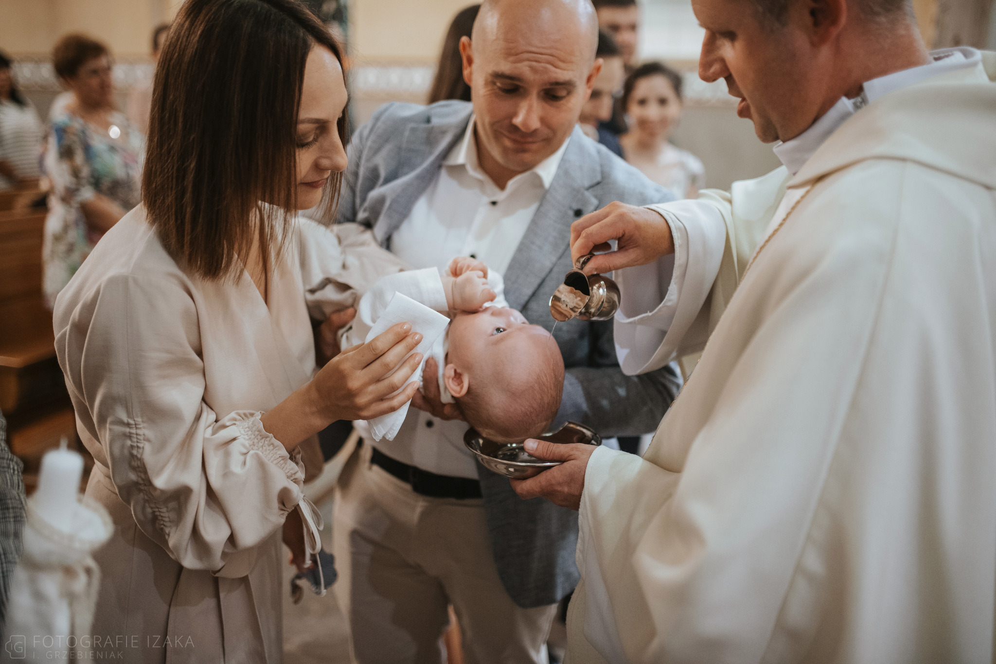 Ksiądz polewa wodą główkę niemowlęcia podczas chrztu, rodzice trzymają dziecko, w tle widoczni goście.