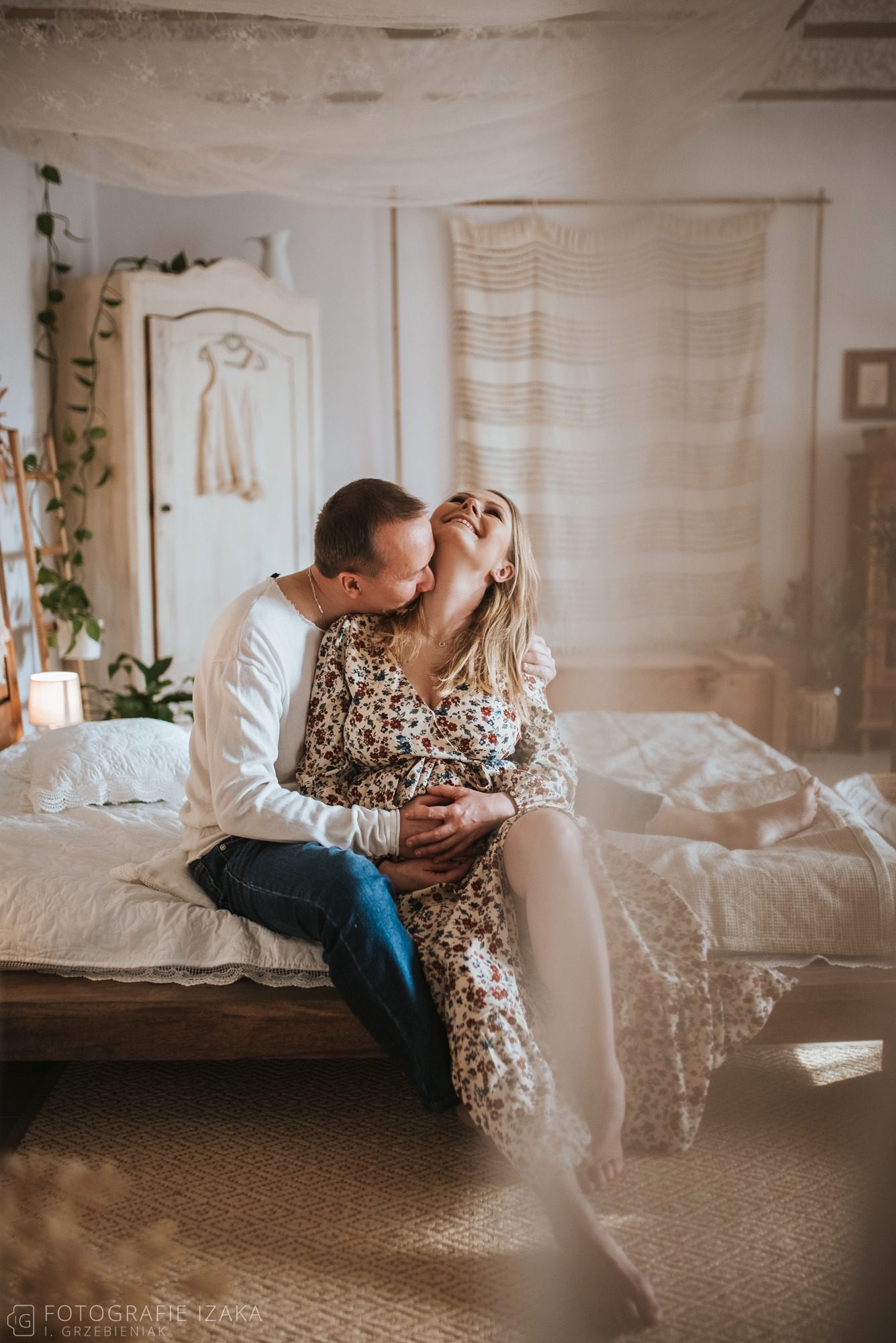 Czułe ujęcie kobiety w kwiecistej sukience w ciąży, obejmowanej przez mężczyznę na łóżku w jasnym, rustykalnym wnętrzu.