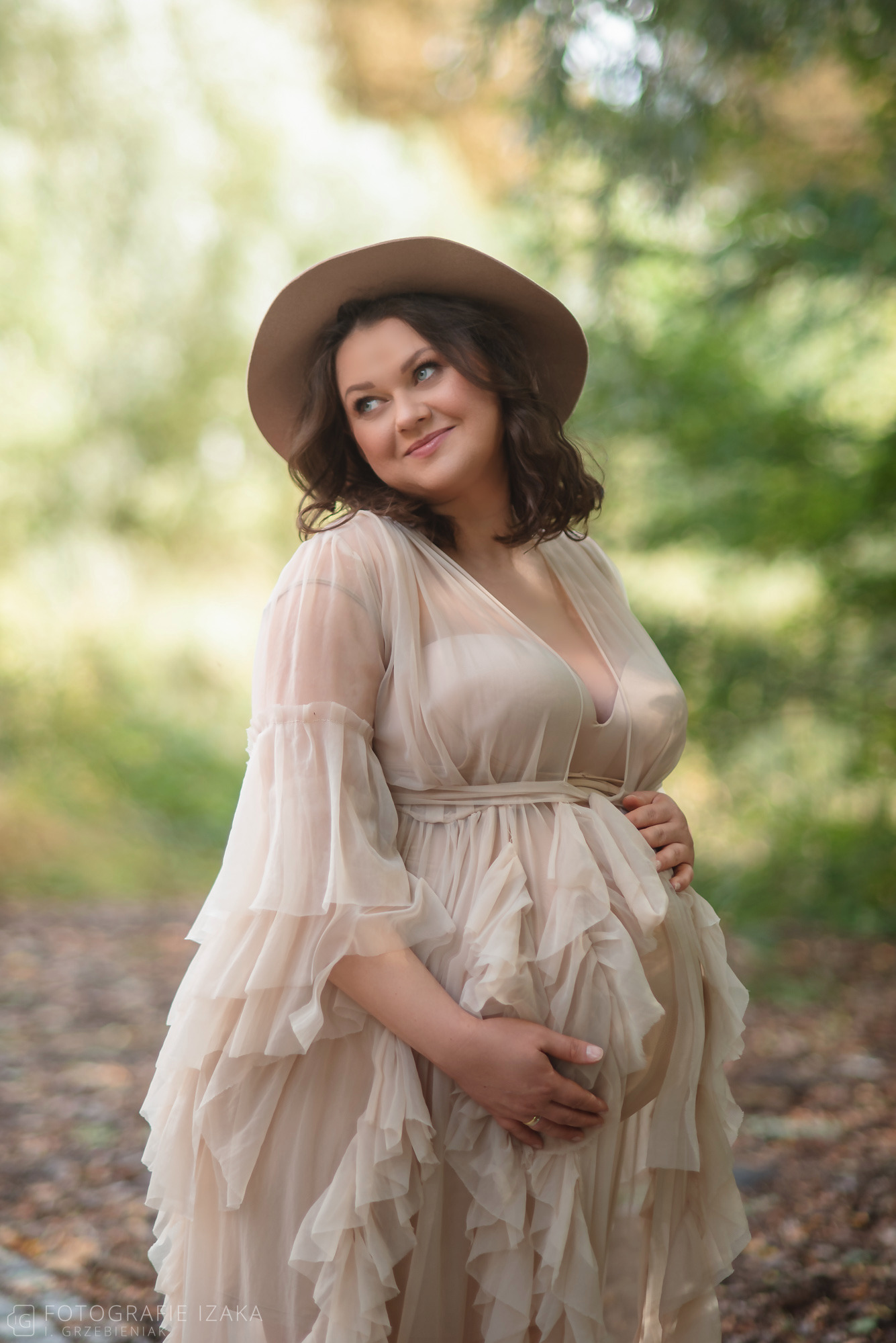Portret kobiety w ciąży w zwiewnej sukience z falbanami i kapeluszu, z rękami na brzuchu, zamyślona poza w plenerze.