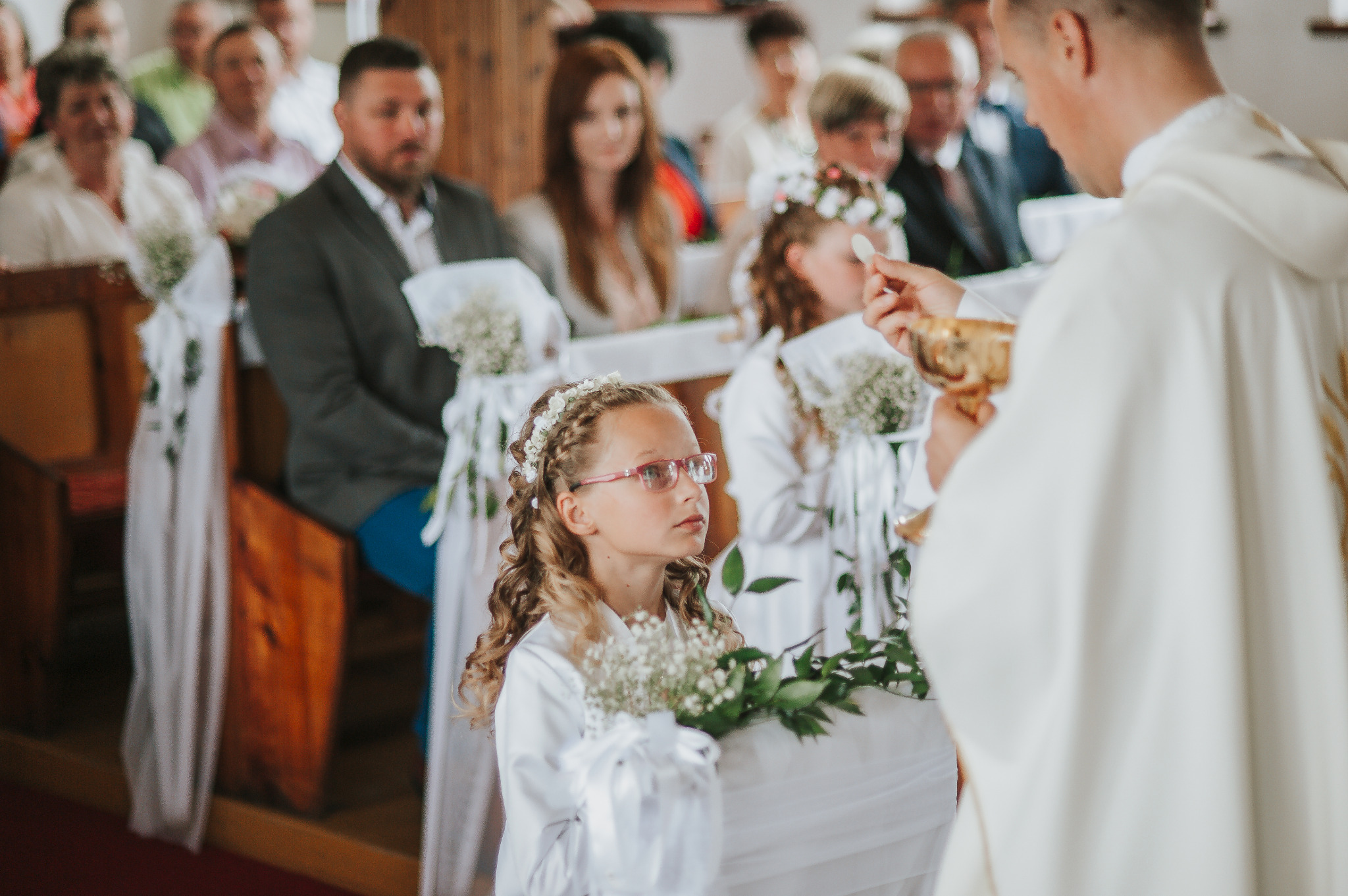 Dziewczynka w okularach podczas Pierwszej Komunii Świętej, trzyma bukiecik, w tle ksiądz podający hostię, widoczni goście w ławkach.