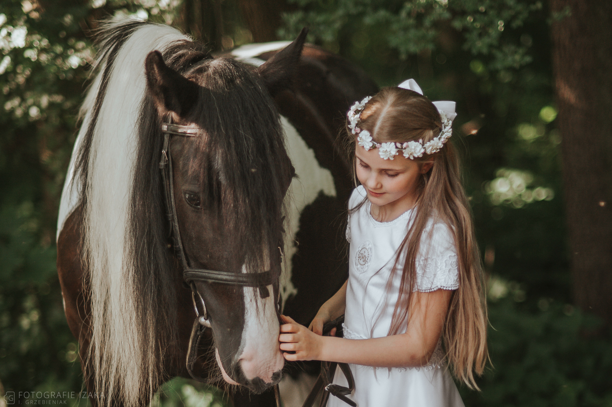 Dziewczynka w białej sukience i wianku głaszcze pysk ciemnego konia z białą grzywą na tle leśnym.
