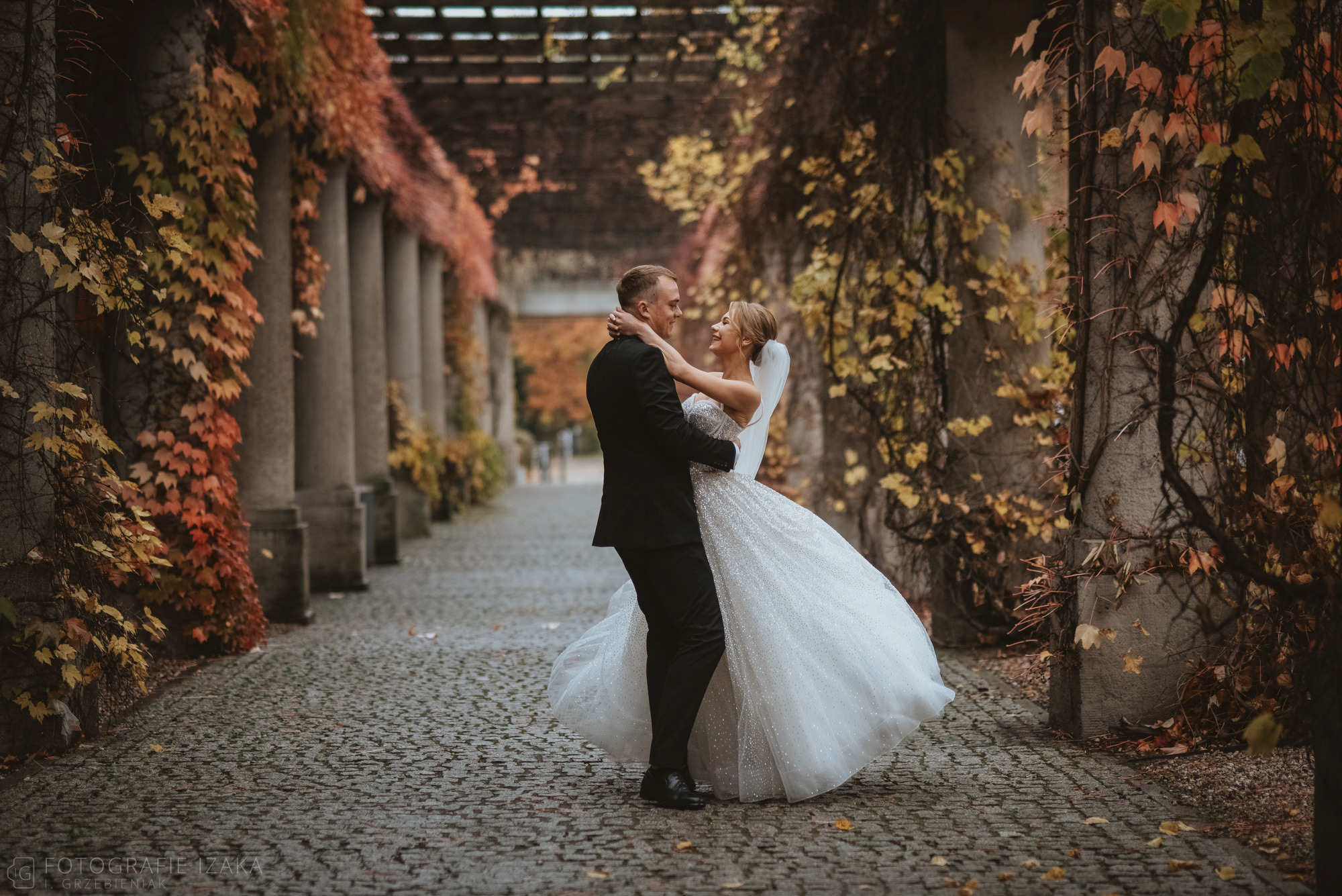 Para młoda obejmuje się w alei z kolumnami porośniętymi bluszczem o jesiennych barwach, panna młoda w błyszczącej sukni i welonie, pan młody w czarnym garniturze.