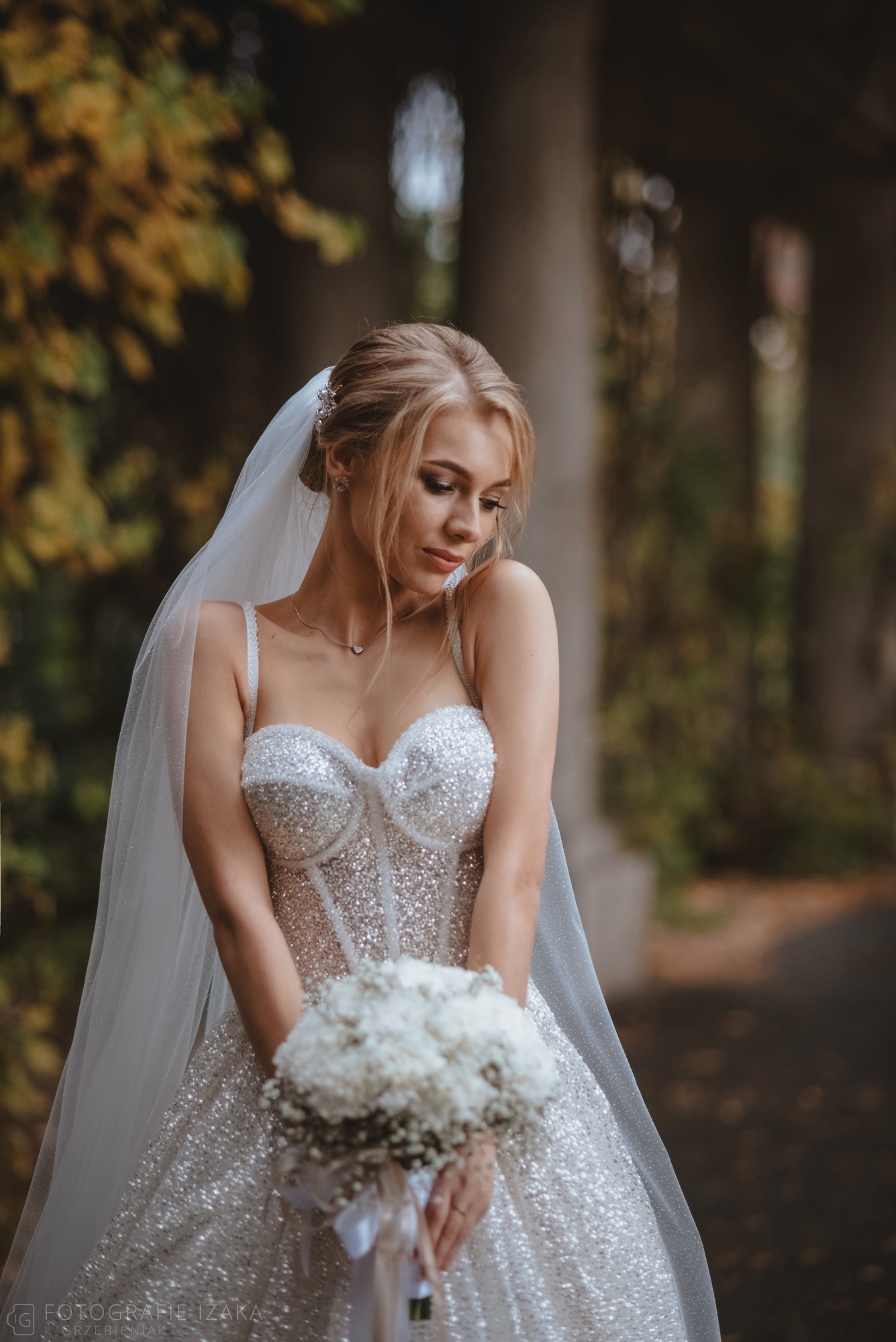 Panna młoda w błyszczącej sukni trzyma bukiet białych kwiatów, spoglądając w dół, z kolumnadą w tle.