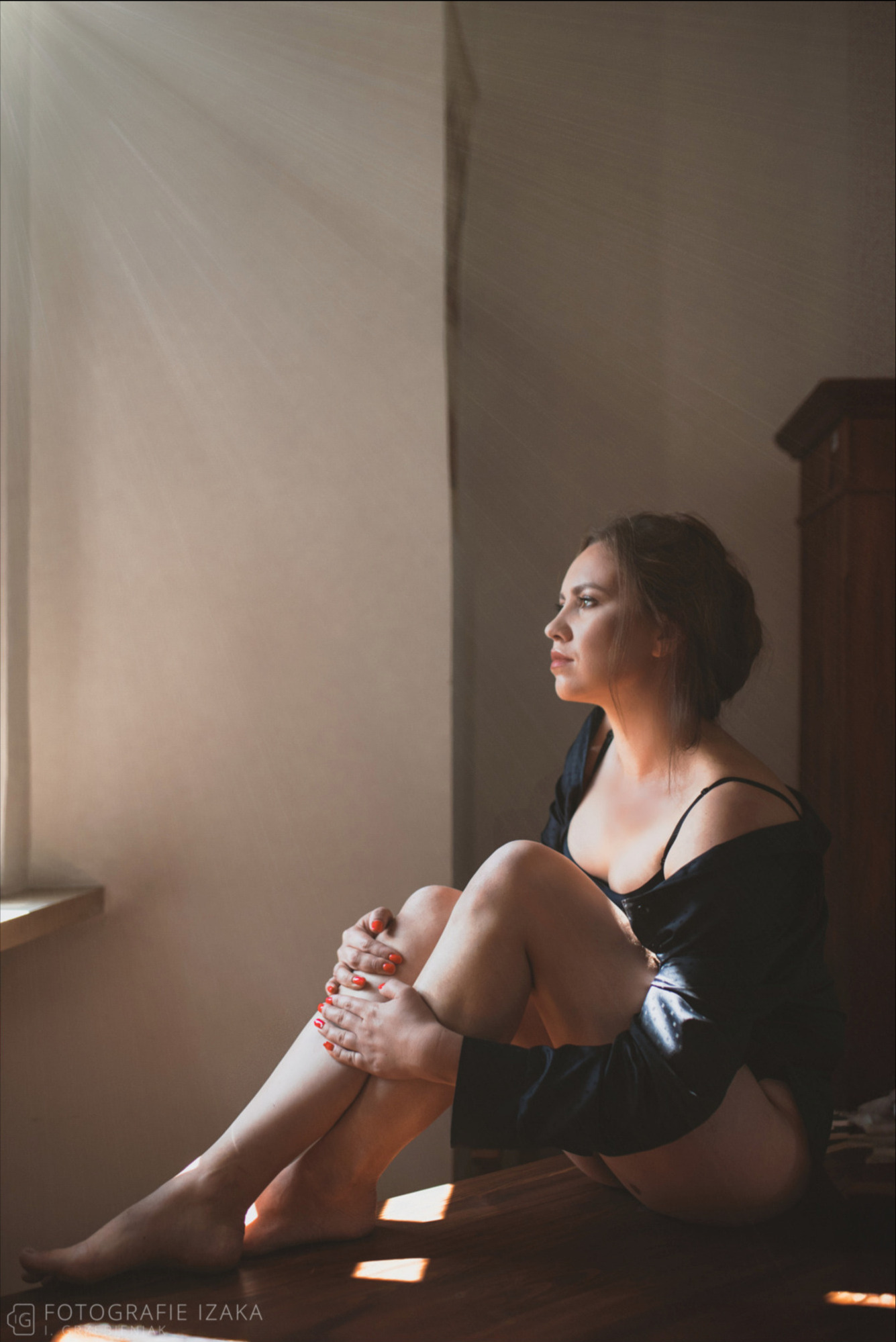 Kobieta siedzi na podłodze w pokoju, spogląda w dal, oświetlona promieniami słońca wpadającymi przez okno, z pomalowanymi na czerwono paznokciami.