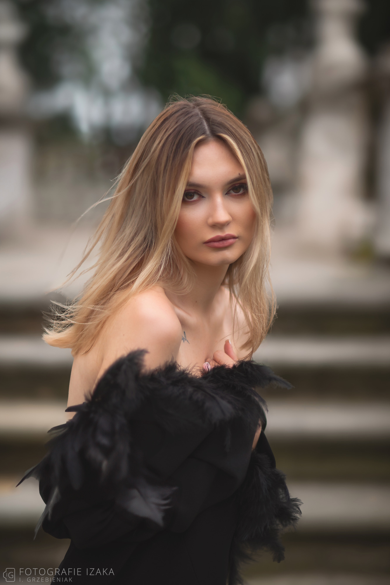 Portret kobiety z blond włosami, owiniętej w czarne pióra, z delikatnym tatuażem na ramieniu, na rozmytym tle schodów.
