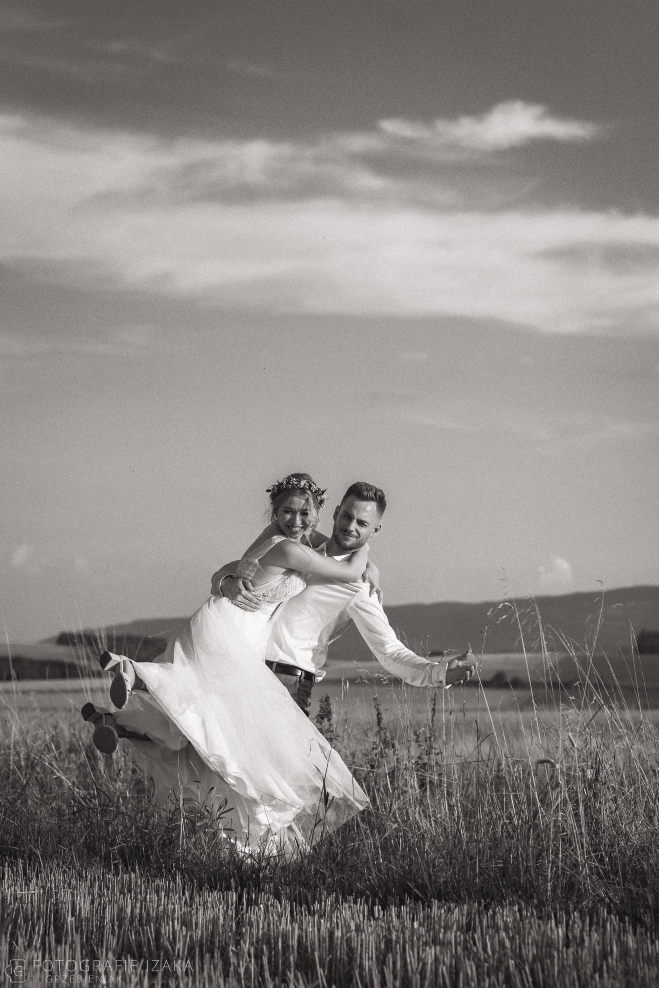 Czarno-białe zdjęcie uśmiechniętej panny młodej w koronie z kwiatów niesionej na barana przez uśmiechniętego pana młodego na tle pola i pochmurnego nieba.