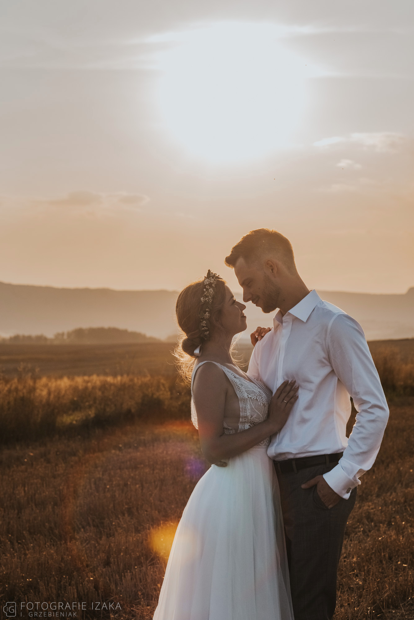 Para młoda w objęciach na tle pola i zachodzącego słońca, panna młoda w sukni z koronkową górą i wiankiem, pan młody w białej koszuli z podwiniętymi rękawami.