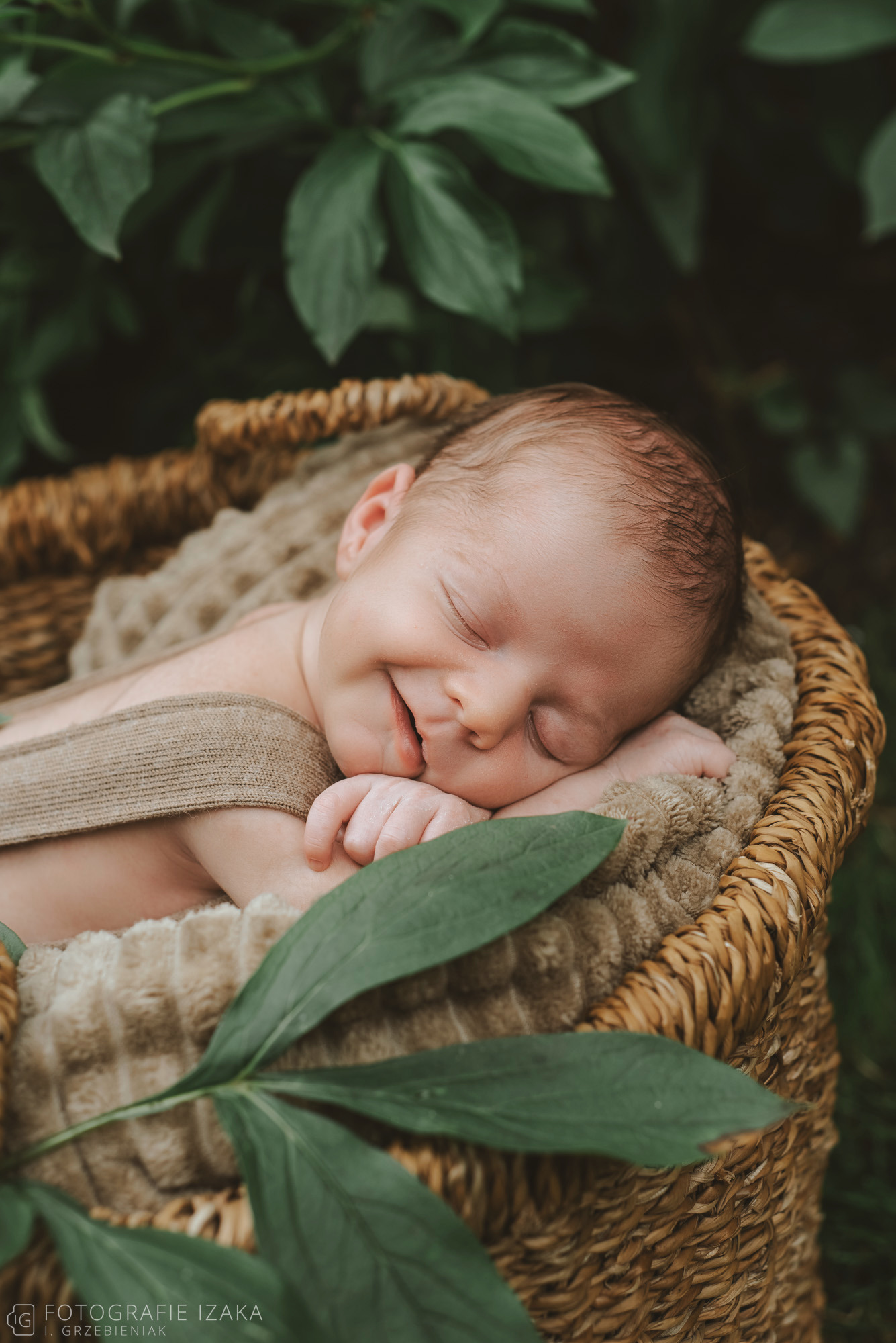 Noworodek śpiący w wiklinowym koszu, otoczony zielonymi liśćmi, z delikatnym uśmiechem na twarzy.