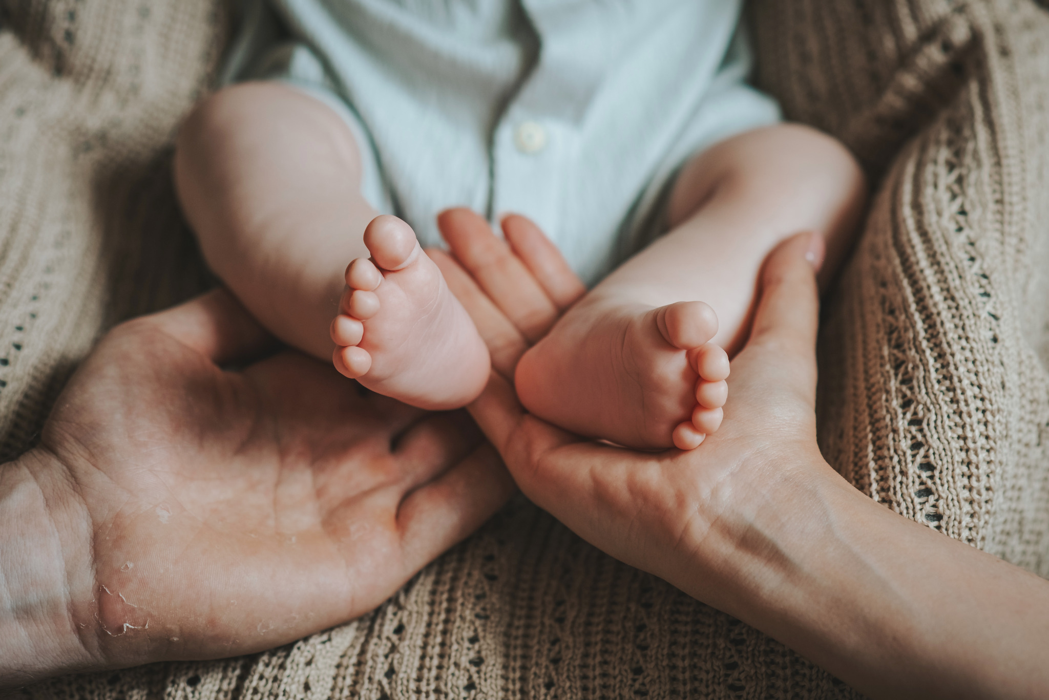 Drobne stópki noworodka spoczywają w dłoniach rodzica na tle beżowej, dzierganej tkaniny. Niewielkie paluszki dziecka, kontrastują z delikatną skórą dłoni dorosłego.