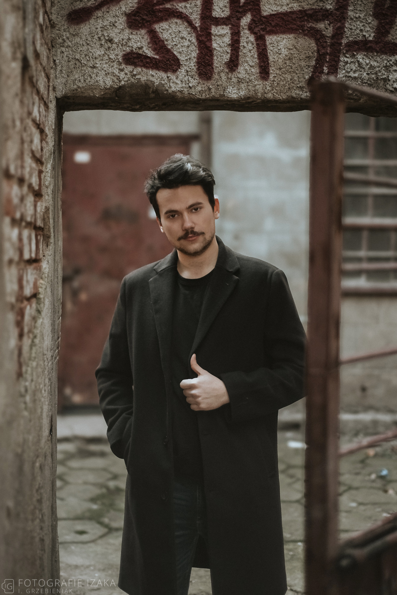 Mężczyzna w ciemnym płaszczu pozuje w przejściu z ceglanymi ścianami i graffiti, dłoń w kieszeni, druga przy klapie, spojrzenie w obiektyw.