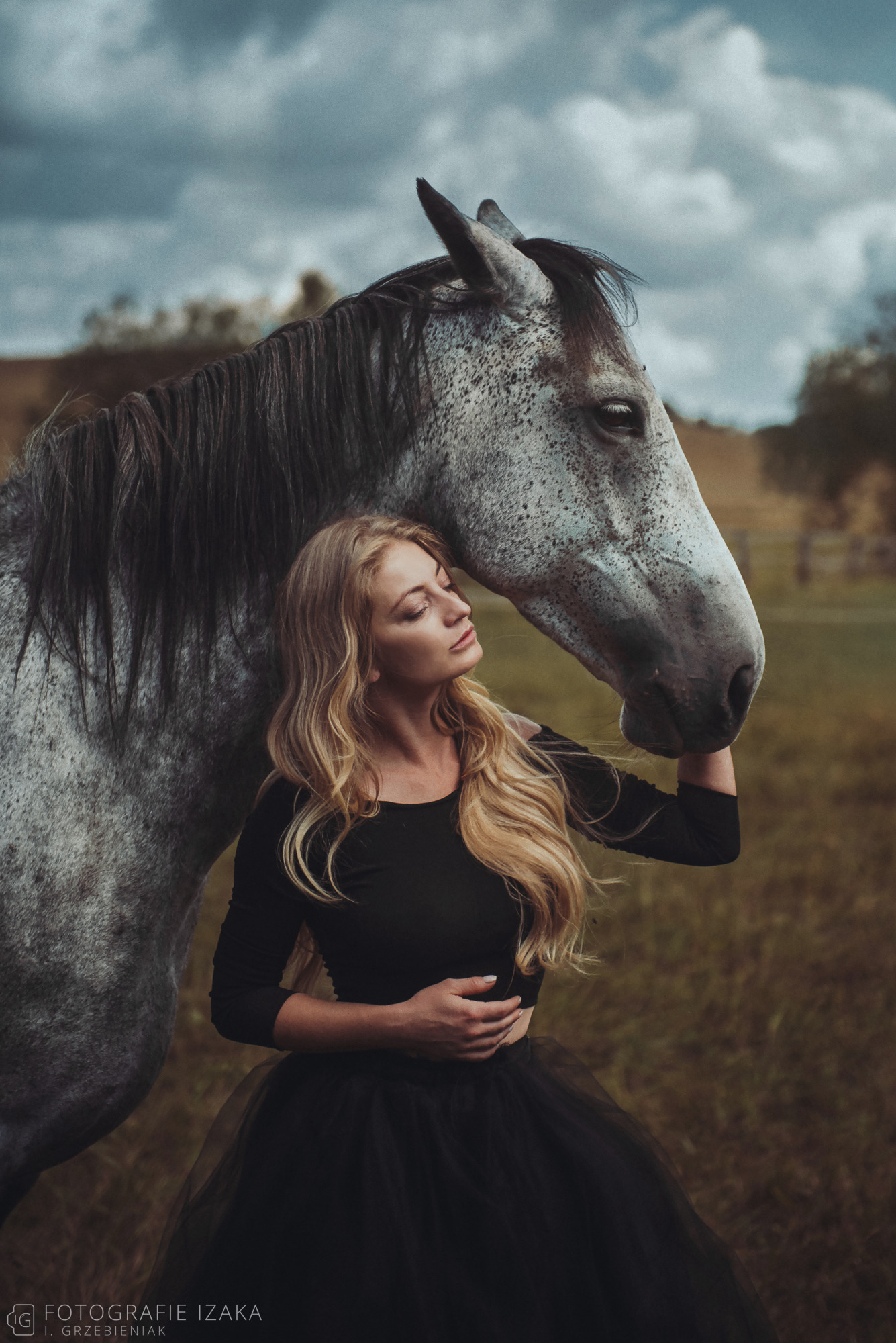 Portret kobiety w czarnej sukni, z zamkniętymi oczami, opierającej policzek o głowę siwego konia na tle łąki i pochmurnego nieba.