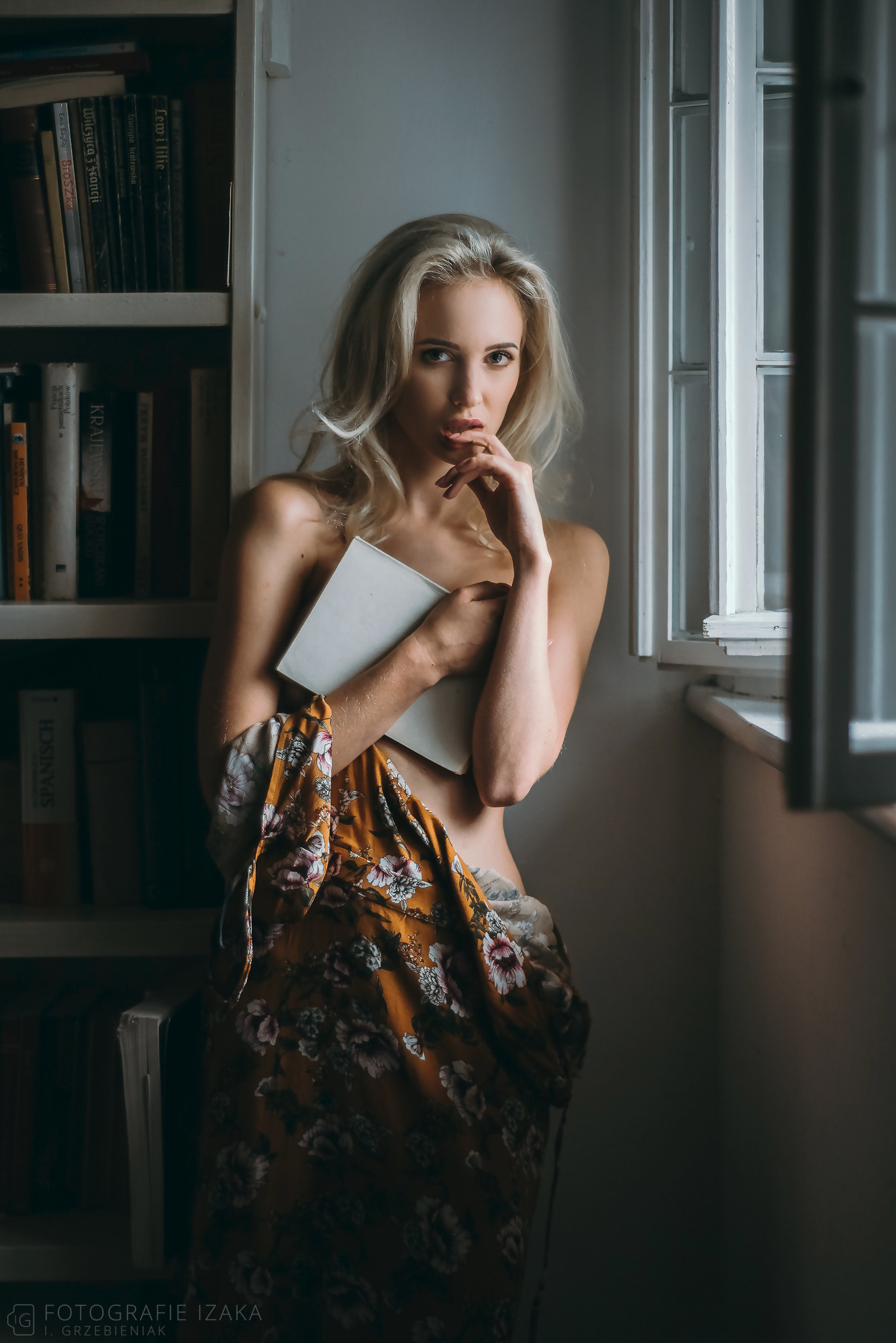 Portret kobiety w stylizacji buduarowej, trzymającej książkę, z fragmentem kwiecistej narzuty, pozującej przy oknie i biblioteczce.