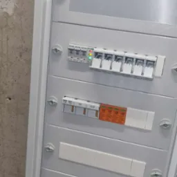 Biała skrzynka rozdzielcza z zainstalowanymi bezpiecznikami, wyłącznikiem głównym Eaton i dwoma licznikami Apator, zamontowana na szarej ścianie.