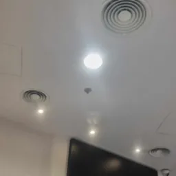 Biały sufit z okrągłymi kratkami wentylacyjnymi, punktowymi lampami LED, czujnikiem dymu i kamerą monitoringu.