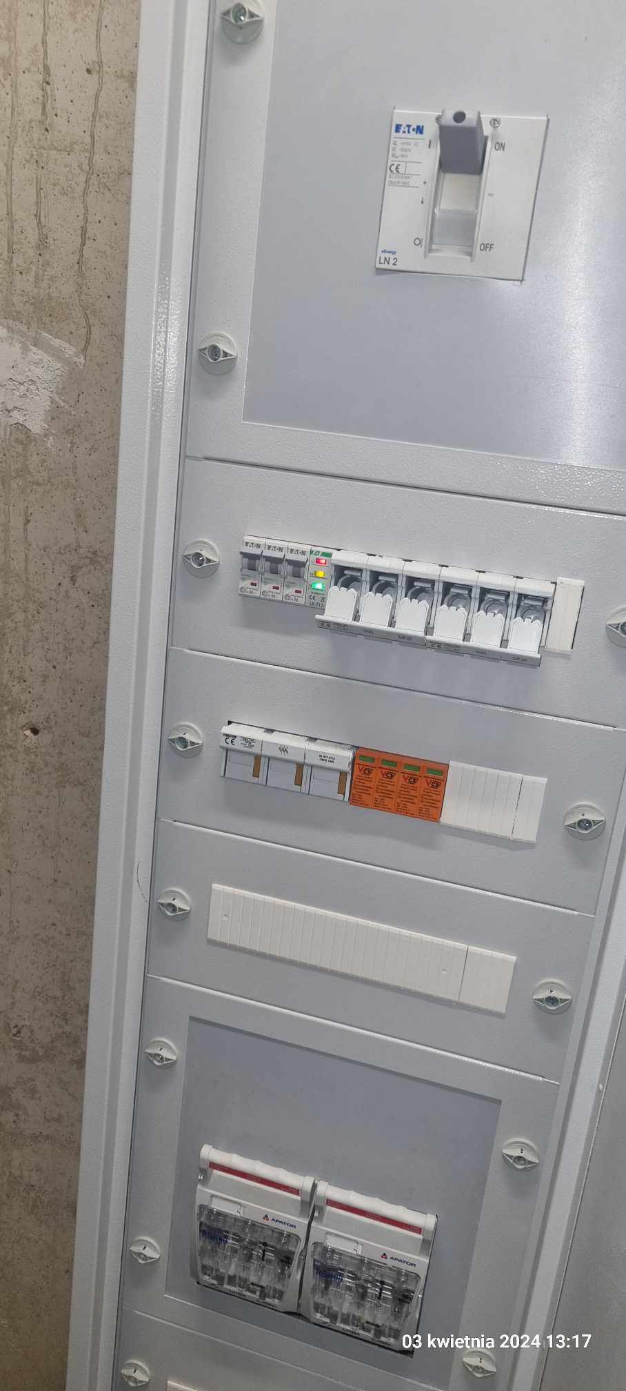 Biała skrzynka rozdzielcza z zainstalowanymi bezpiecznikami, wyłącznikiem głównym Eaton i dwoma licznikami Apator, zamontowana na szarej ścianie.