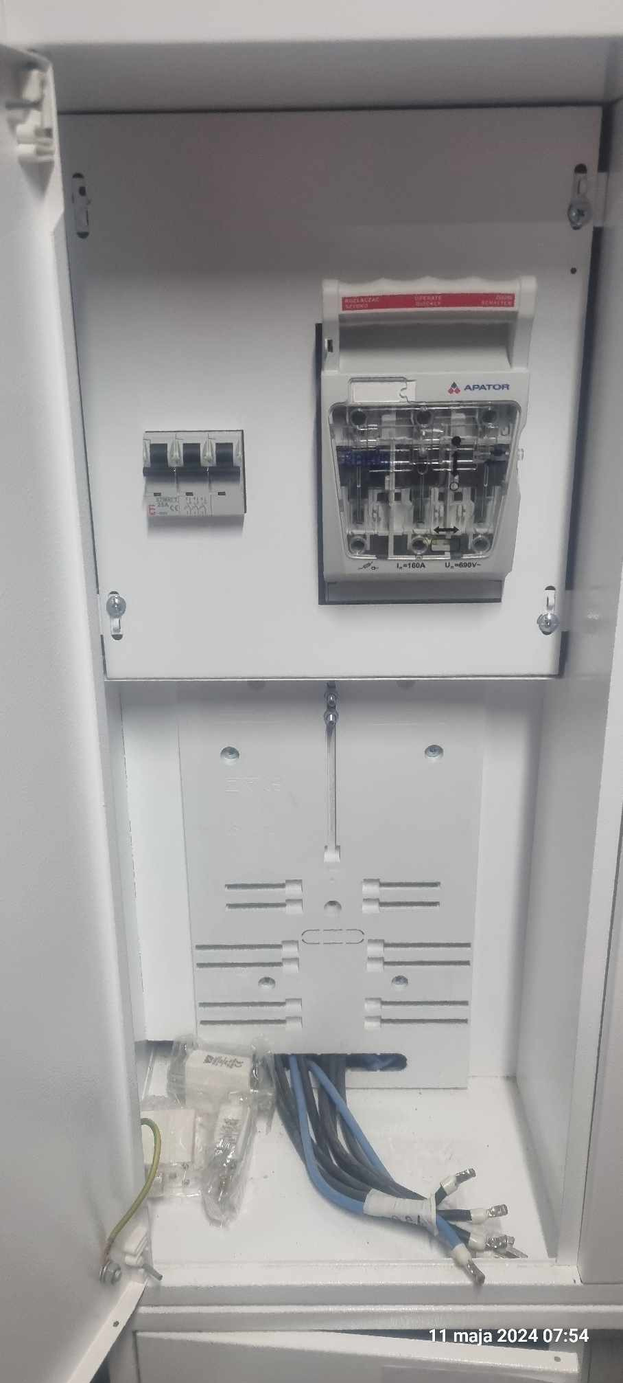 Wnętrze białej skrzynki elektrycznej z zamontowanym bezpiecznikiem trójfazowym, licznikiem energii marki Apator i przygotowanymi przewodami z końcówkami.
