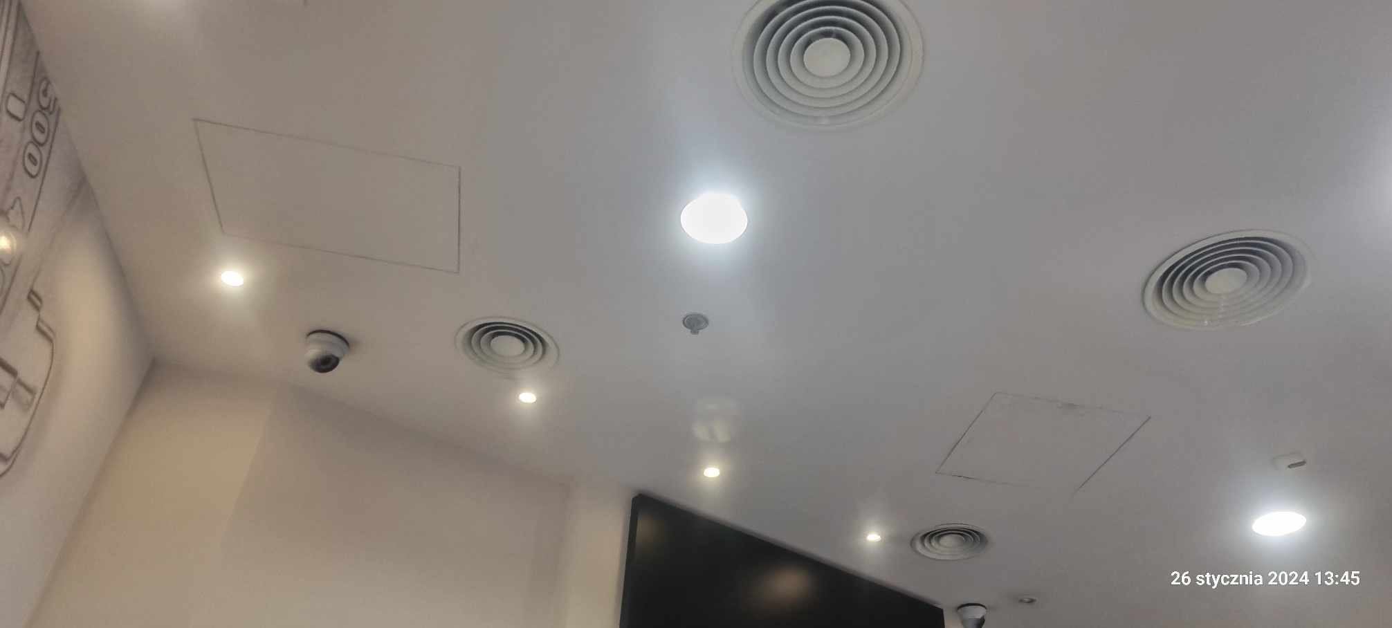 Biały sufit z okrągłymi kratkami wentylacyjnymi, punktowymi lampami LED, czujnikiem dymu i kamerą monitoringu.