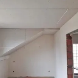 Wnętrze pokoju w trakcie montażu płyt kartonowo-gipsowych na suficie skośnym i ścianach, widoczne profile i śruby mocujące, surowy otw&oacute;r drzwiowy z ceglaną obudową w tle.