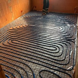 SantechPro - Ułożone, niebieskie rury ogrzewania podłogowego na folii, widoczne fragmenty ścian z cegły dziurawki i instalacja elektryczna. Częściowo widoczna osoba w roboczym ubraniu.