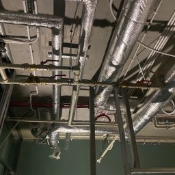 SantechPro - Instalacja rur i przewodów pod sufitem w trakcie remontu. Widoczne rury izolowane, rury PCV, przewody elektryczne i metalowa konstrukcja szkieletowa.