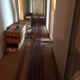 SantechPro - Instalacja grzewcza podłogowa w korytarzu w trakcie montażu. Widoczne rury w kolorze czerwonym i niebieskim, prowadzone wzdłuż ścian. Na podłodze kartony z materiałami.